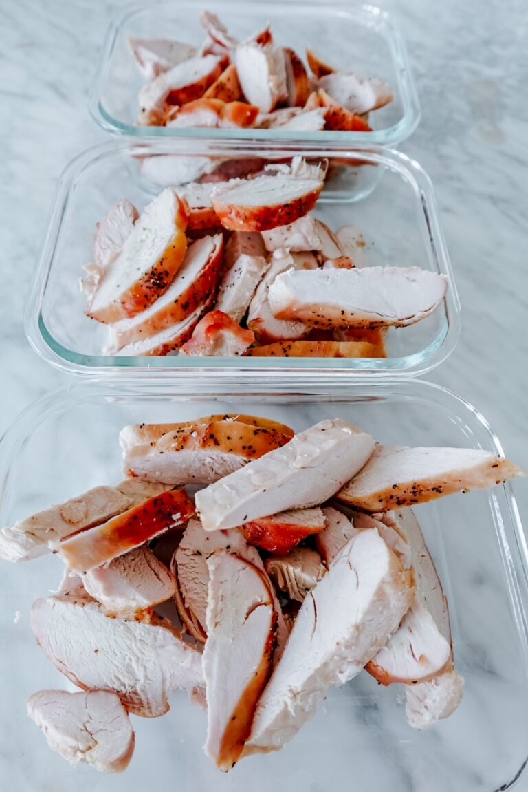 Three glass meal prep containers filled with sliced, cooked chicken breast on a marble surface.