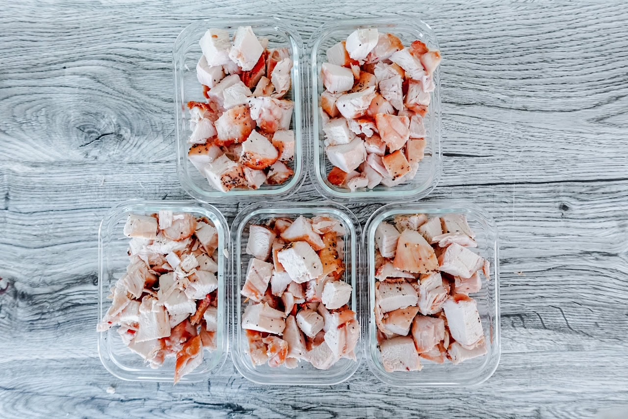 Five glass containers filled with diced chicken arranged neatly on a wooden surface, ready for meal prep or freezing.