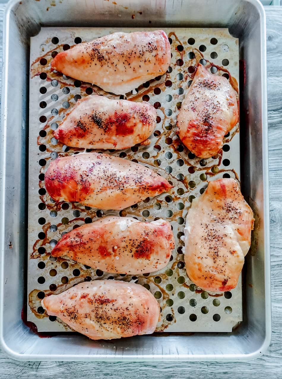 Overhead view of cooked chicken breasts on a metal tray with holes sitting inside a metal pan.