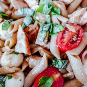 A detailed close-up of Caprese chicken pasta with juicy tomatoes, chunks of chicken, mozzarella pearls, and chopped basil tossed together.