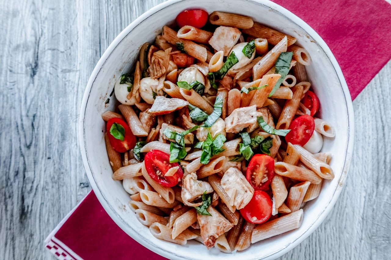 A white bowl filled with penne pasta, grilled chicken, cherry tomatoes, mozzarella balls, and fresh basil leaves, set on a wooden table with a red napkin.