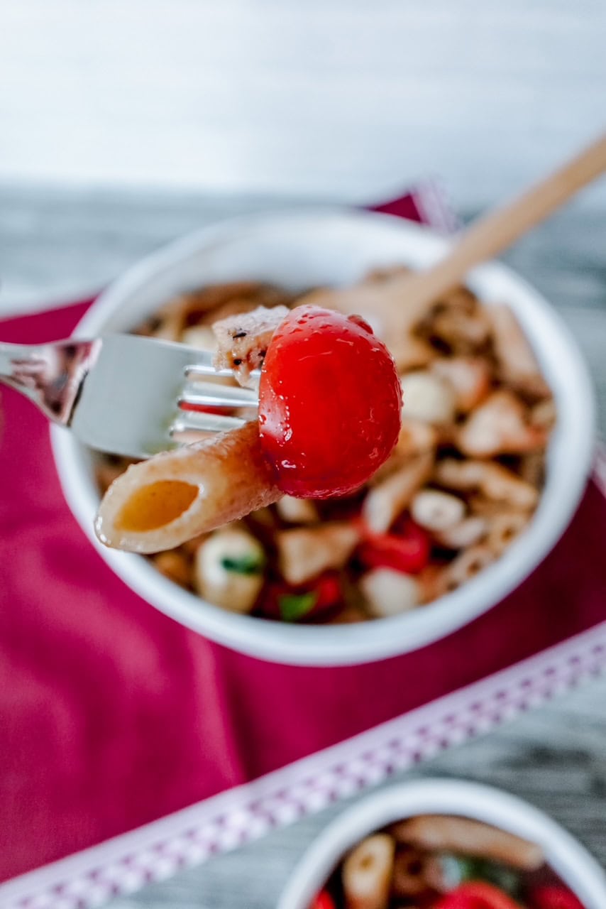 A close-up of a fork holding a cherry tomato and piece of pasta above a bowl of Caprese chicken pasta.