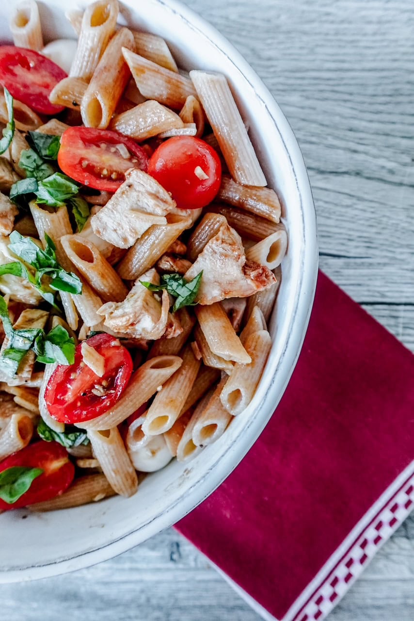 A white bowl filled with penne pasta, grilled chicken, cherry tomatoes, mozzarella balls, and fresh basil leaves, set on a wooden table with a red napkin.