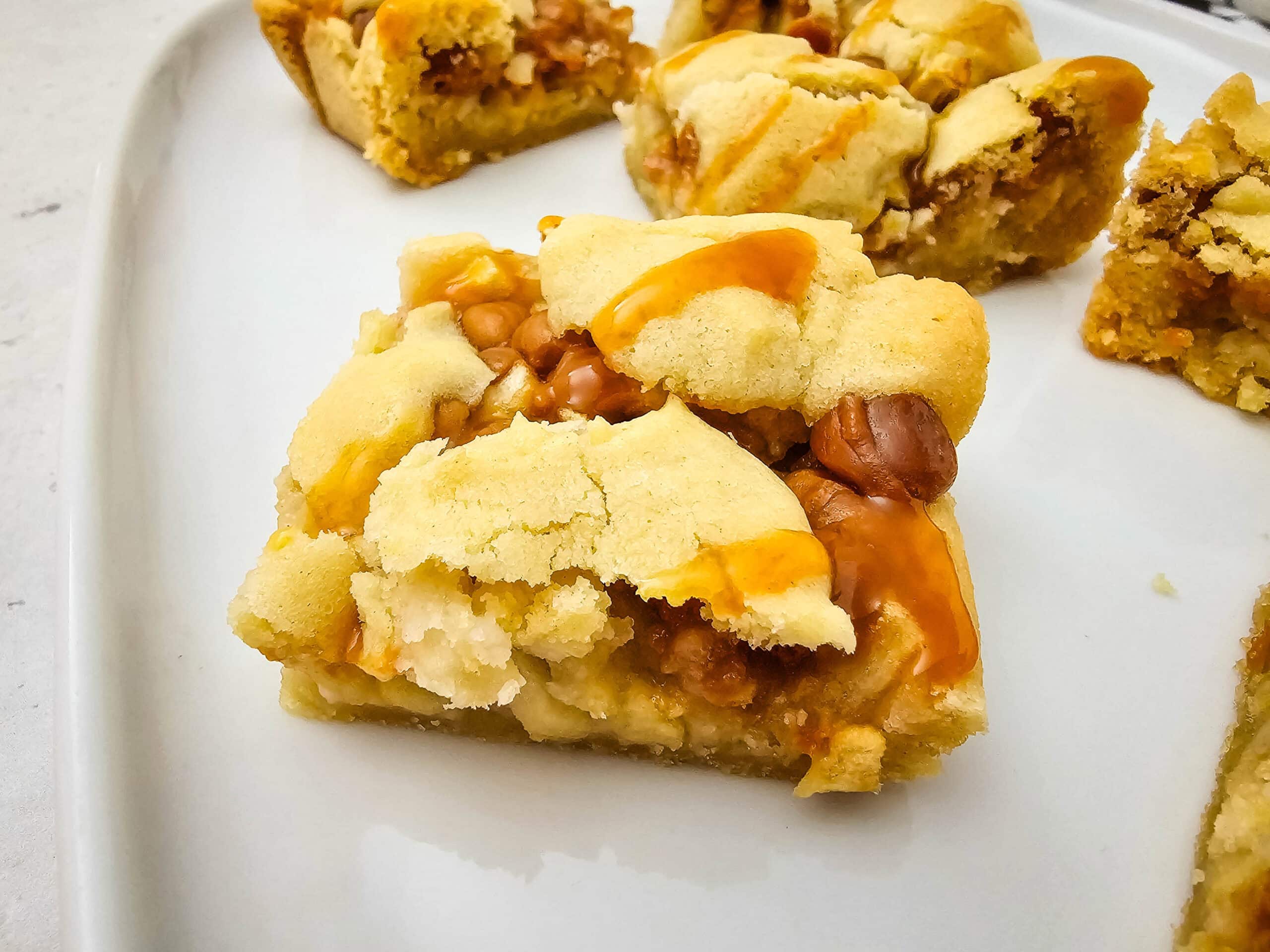 A close-up of a caramel apple cookie bar on a white plate, showing a soft, crumbly texture with visible caramel drizzle and apples. Other bars are blurred in the background.