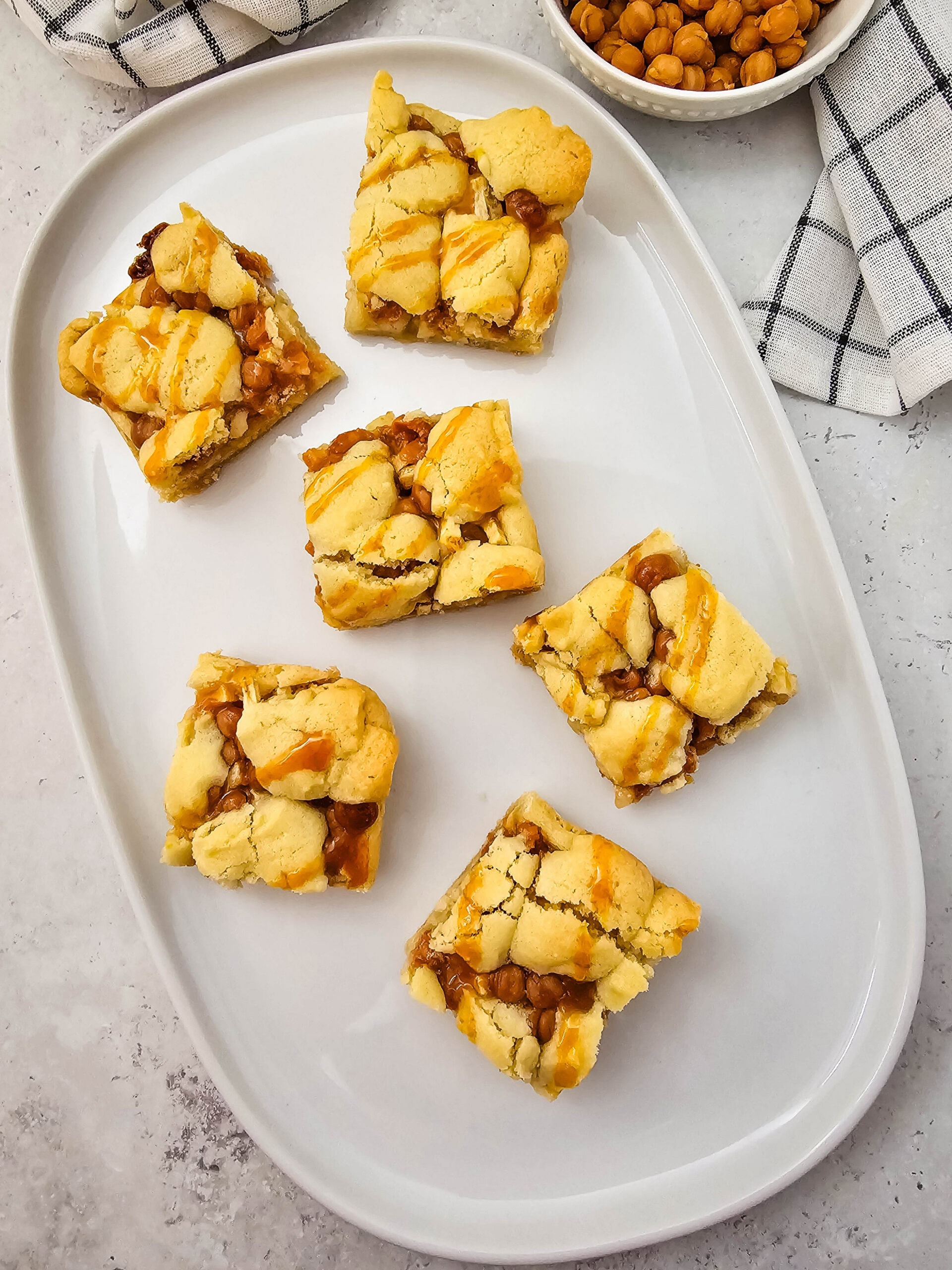 Six caramel apple bars with golden, crumbly tops and caramel chips are arranged on a white oval plate. A bowl of caramel chips and a checkered towel are visible in the background.