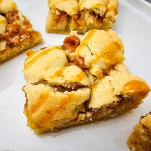 A close-up of caramel apple bars with a golden, crumbly crust and gooey caramel drizzled on top, arranged on a white plate.