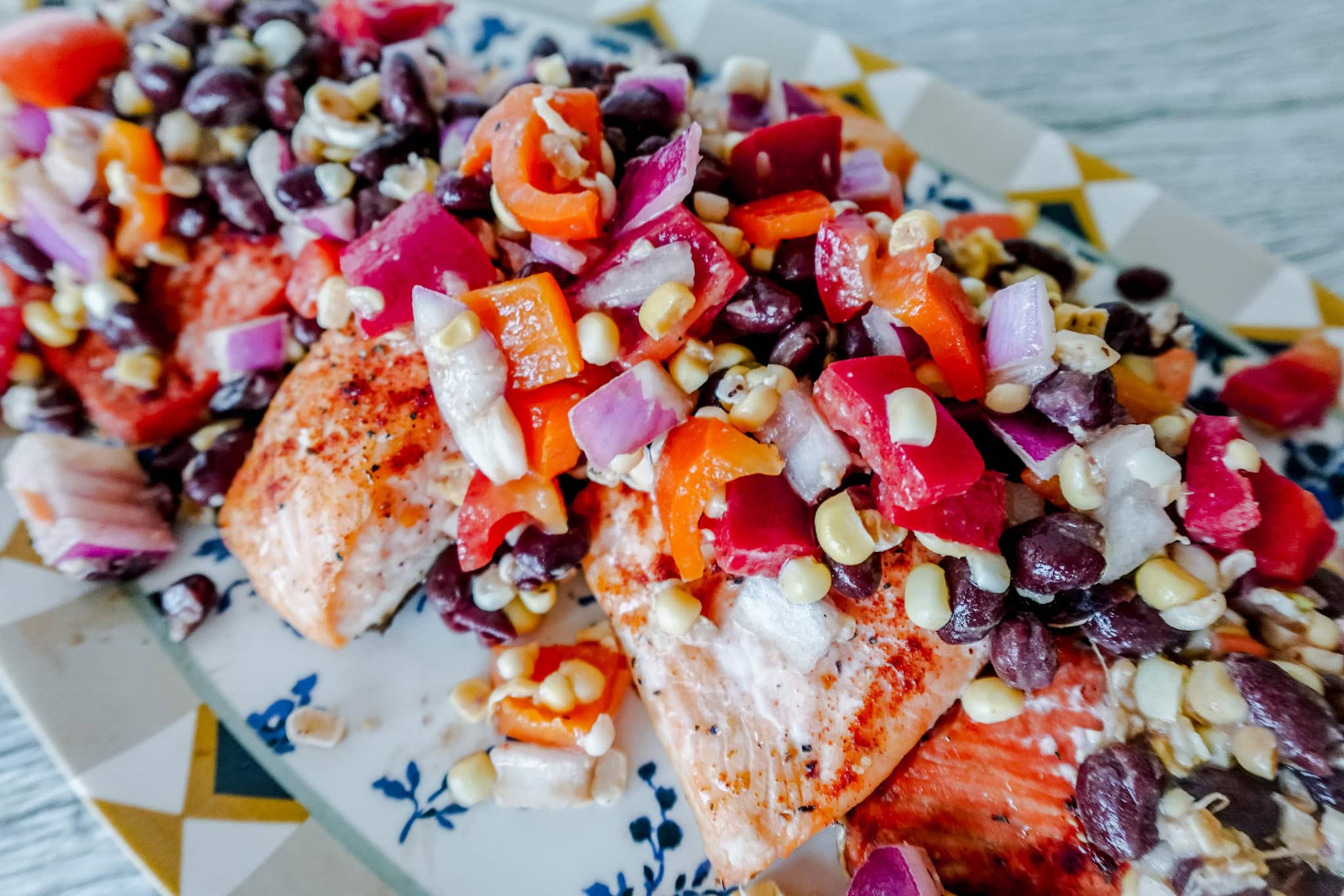 Grilled salmon fillets topped with a colorful mixture of black beans, corn, chopped red onions, and red bell peppers, served on a patterned ceramic platter.