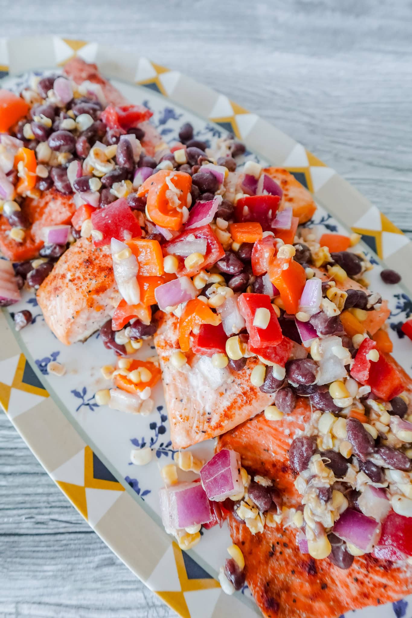 Grilled salmon fillets topped with a colorful mixture of black beans, corn, red onions, tomatoes, and peppers, served on a patterned platter.