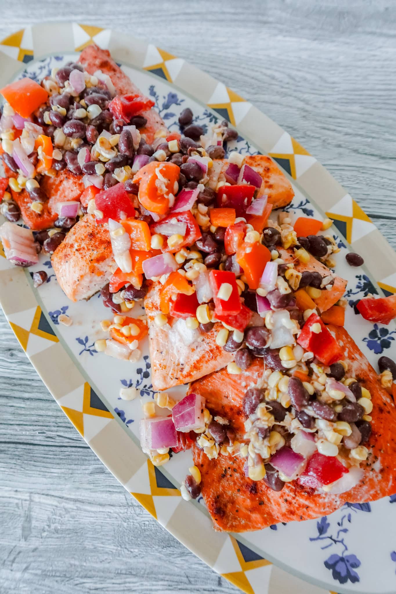 Grilled salmon fillets topped with a colorful mixture of black beans, corn, diced tomatoes, and red onions served on a patterned oval plate.