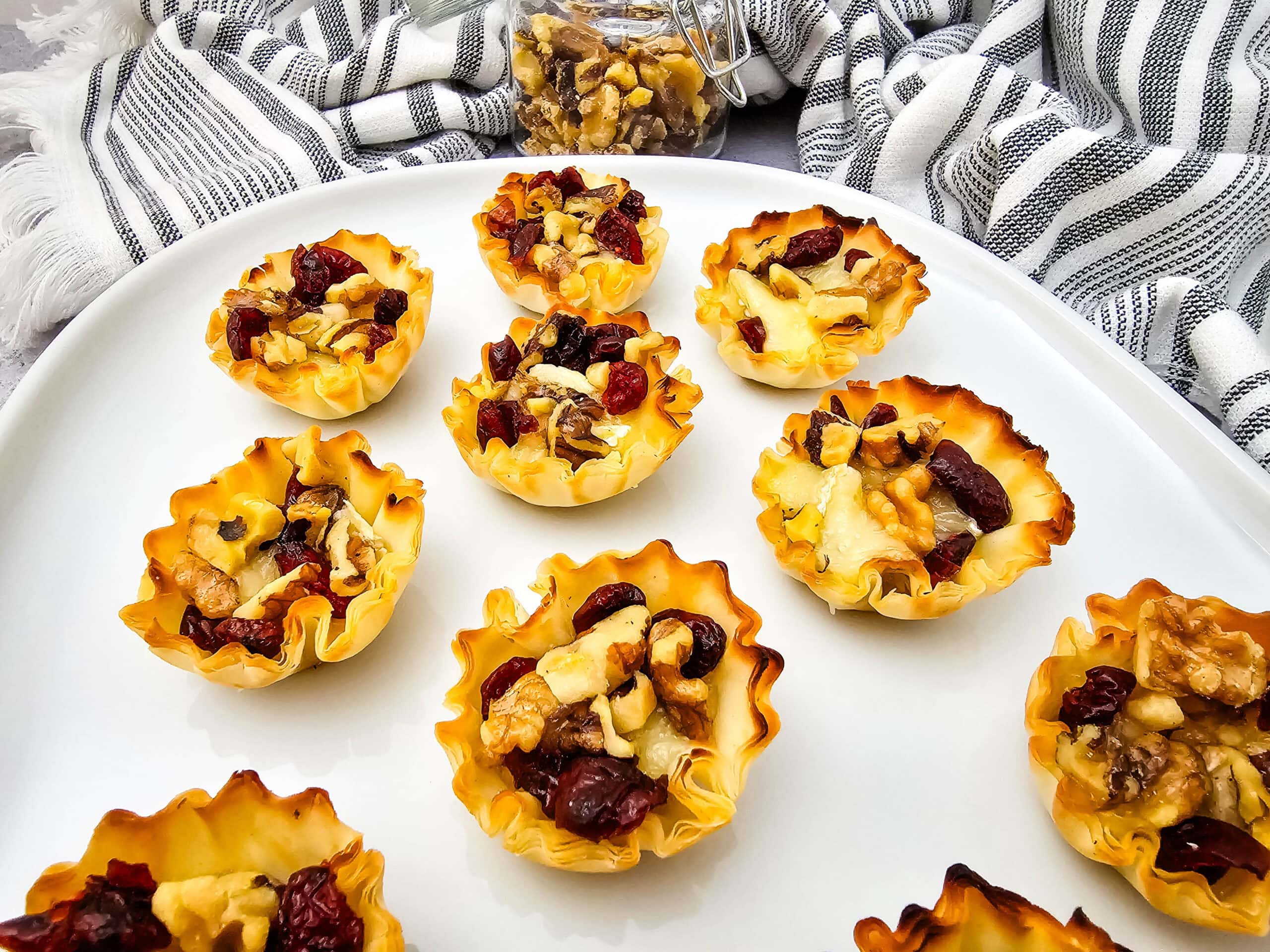 Mini phyllo pastry cups filled with walnuts, dried cranberries, and melted cheese are arranged on a white platter. A jar of walnuts and a striped cloth are in the background.