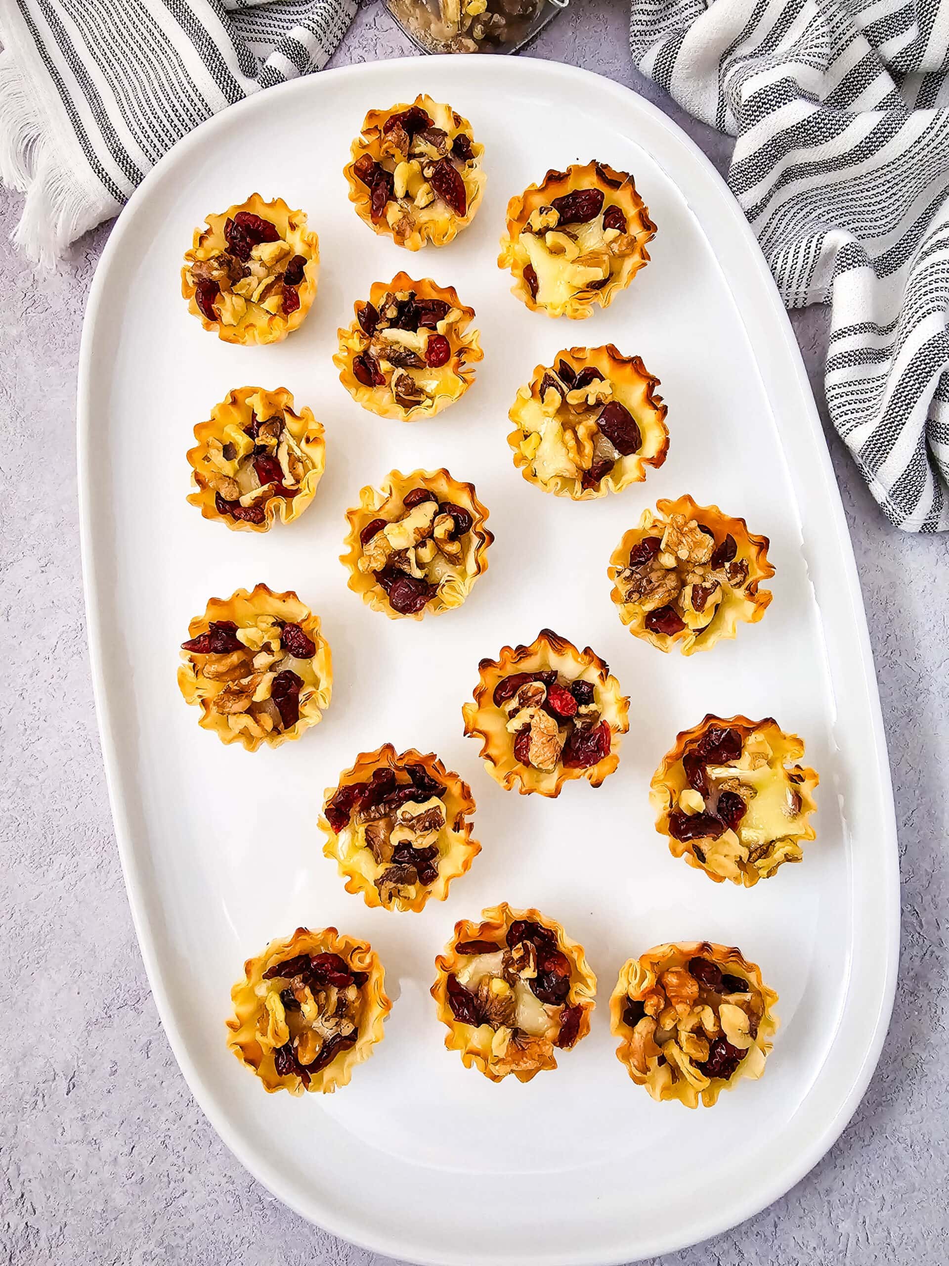 A white platter displays fifteen mini phyllo pastry cups filled with chopped nuts and dried fruits. The platter is set on a light surface with a striped towel partially visible in the background.