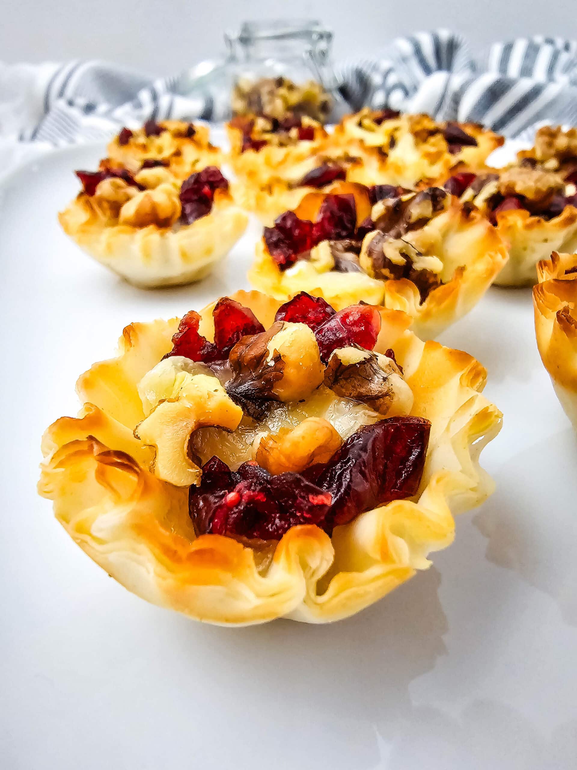 Mini phyllo pastry cups filled with chopped walnuts and dried cranberries, arranged on a white plate, with a striped cloth and a glass jar in the background.