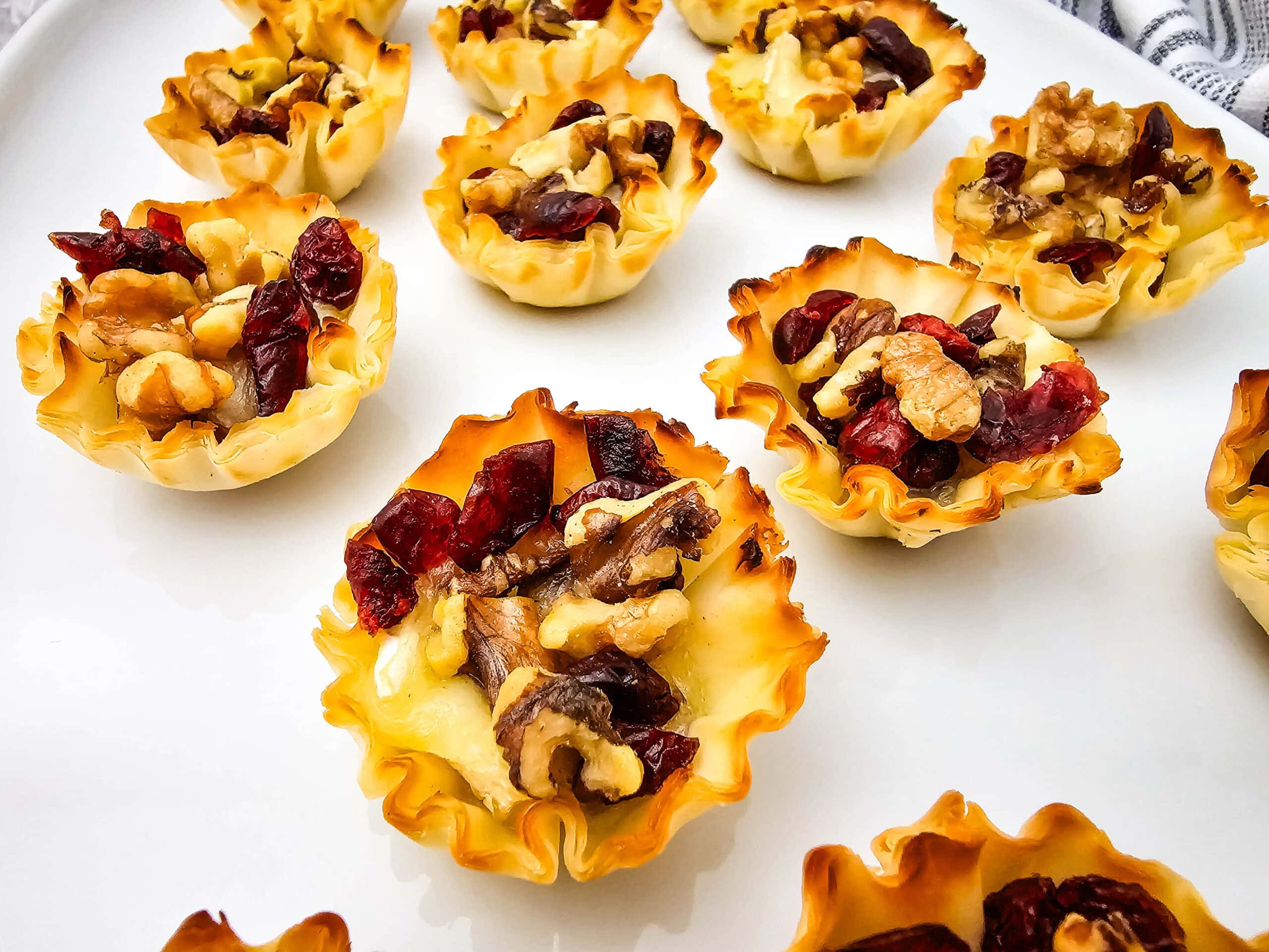 Mini phyllo pastry cups filled with melted cheese, walnuts, and dried cranberries are arranged on a white plate. The cups have golden brown, crisp edges and are displayed in close-up detail.