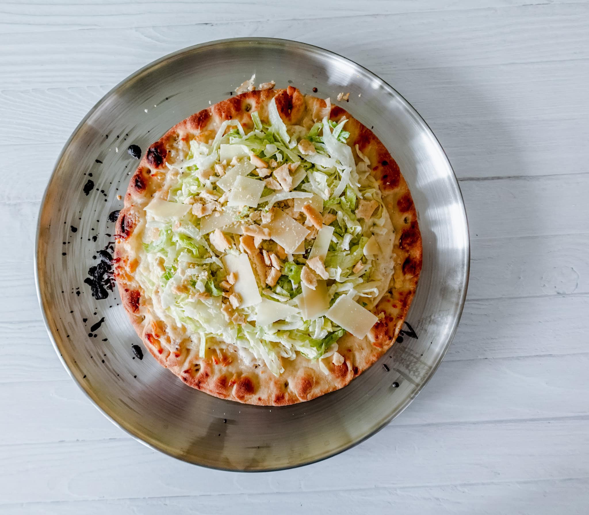 Flatbread pizza topped with shredded lettuce, shaved Parmesan cheese, and crushed croutons on a silver plate.