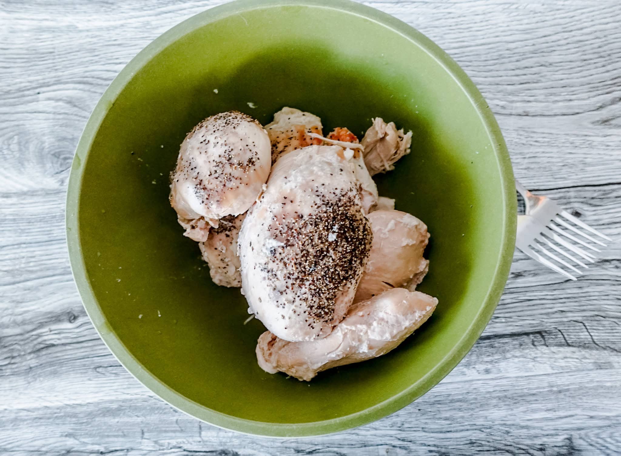 An overhead shot of a green bowl on a gray wooden table with forks next to it. In the bowl is a pile of cooked chicken breasts seasoned with pepper.