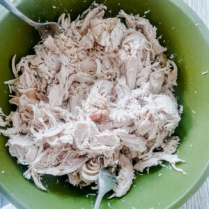 An overhead shot of shredded seasoned chicken in a green bowl with forks setting against the side for shredding. A white cloth peaks out of the lower right corner.