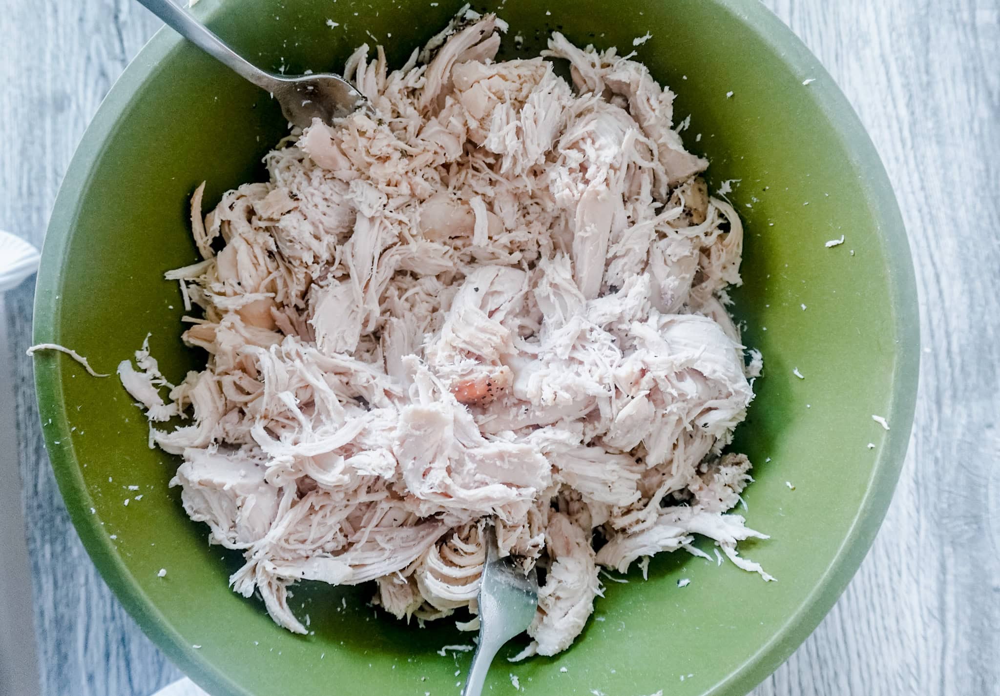 An overhead shot of shredded seasoned chicken in a green bowl with forks setting against the side for shredding. A white cloth peaks out of the lower right corner.
