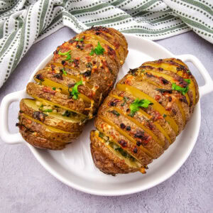 Two baked Hasselback potatoes are topped with crisp herbs and melted cheese, served on a white dish. A green and white striped cloth napkin is framing the top half of the image.