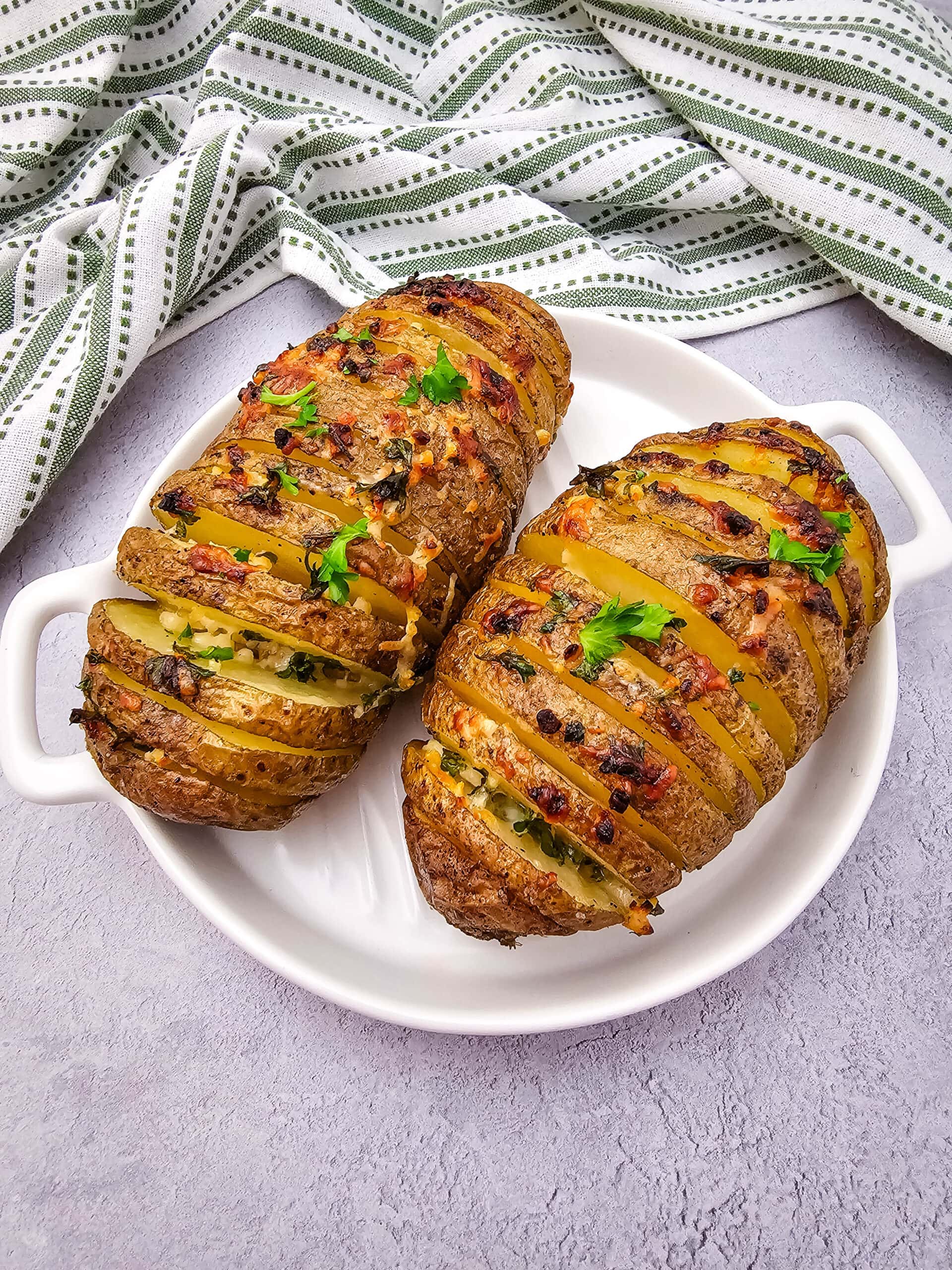 Two baked Hasselback potatoes are topped with crisp herbs and melted cheese, served on a white dish. A green and white striped cloth napkin is framing the top half of the image.