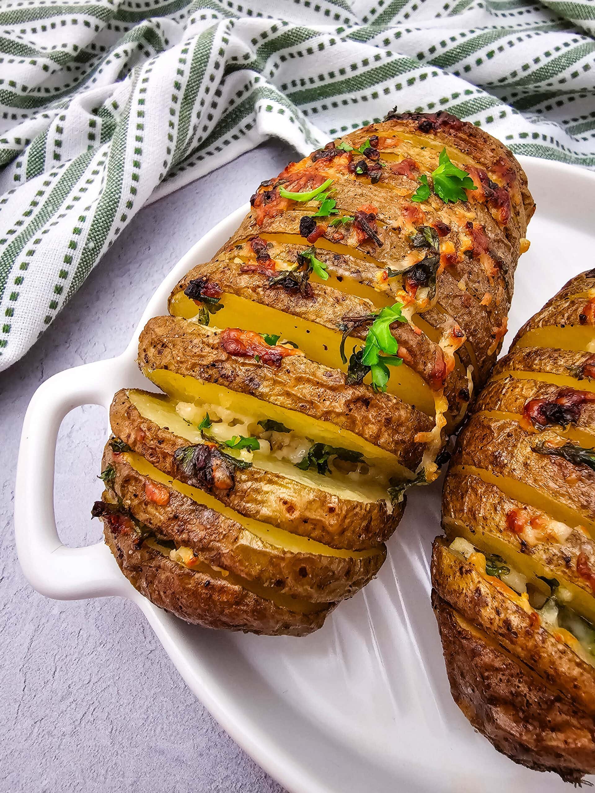 Close-up of two baked Hasselback potatoes on a white dish, topped with melted cheese, herbs, and parsley. A green and white striped cloth lies in the background.