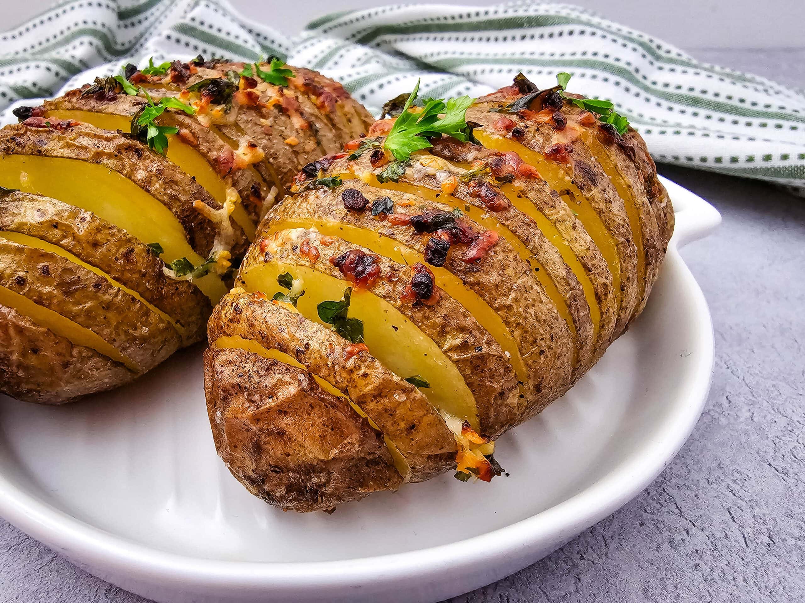 Two golden-brown Hasselback potatoes are on a white plate, garnished with herbs and melted cheese, against a green and white striped towel background.
