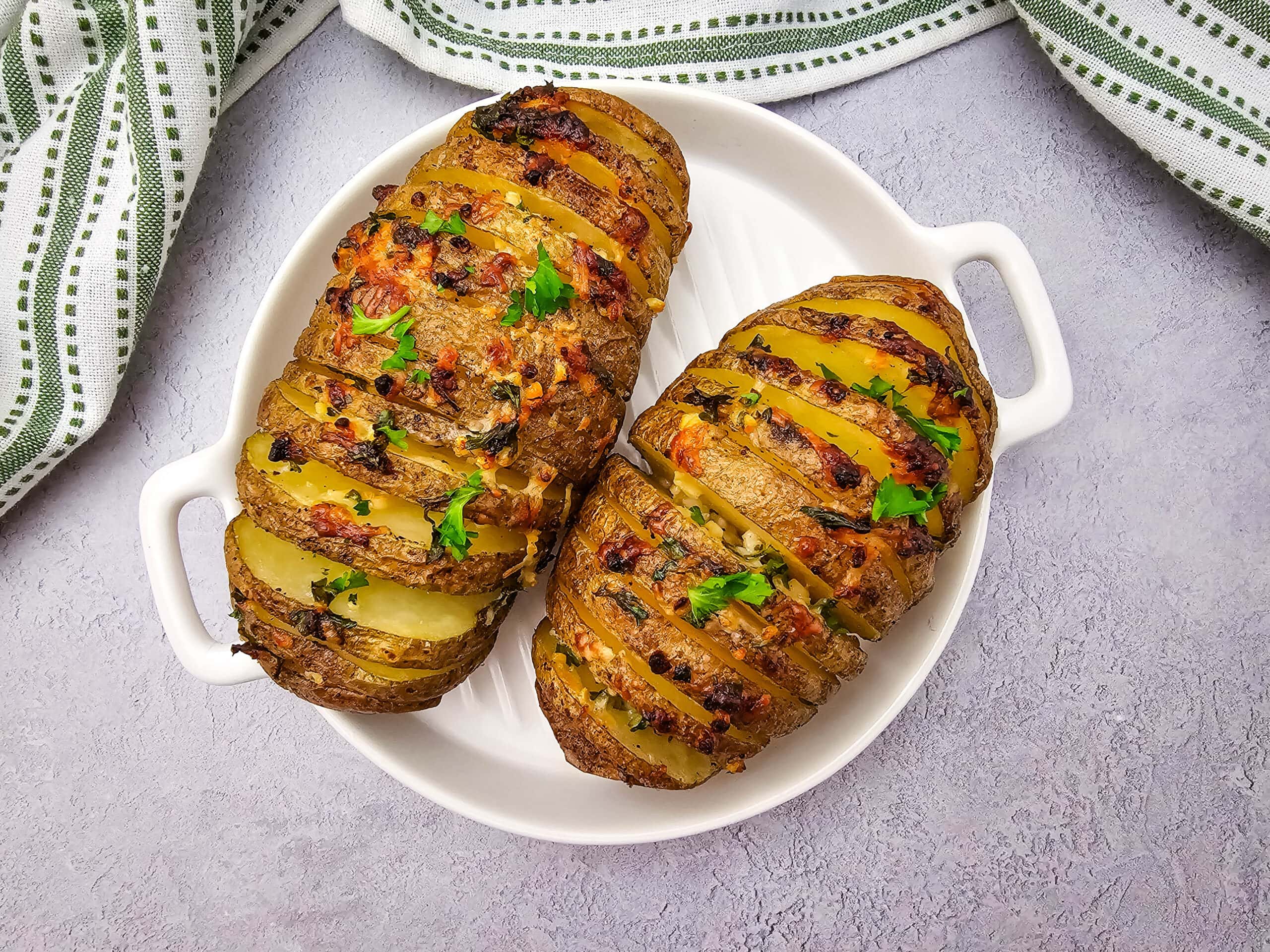 Two Hasselback baked potatoes with herbs and cheese are served on a white circular dish, placed on a light gray surface with a green and white striped kitchen towel nearby.