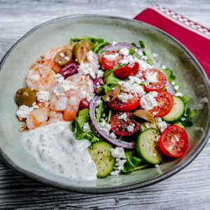 A bowl of salad with shrimp, sliced cucumbers, cherry tomatoes, red onions, olives, crumbled feta cheese, greens, and a serving of creamy white dressing, placed on a wooden surface beside a red napkin.