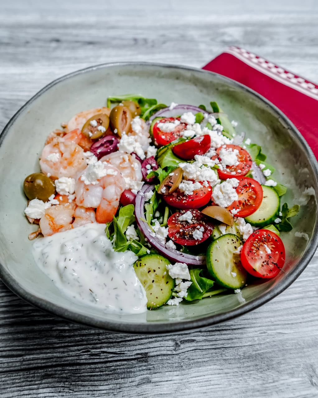 A bowl of salad with shrimp, sliced cucumbers, cherry tomatoes, red onions, olives, crumbled feta cheese, greens, and a serving of creamy white dressing, placed on a wooden surface beside a red napkin.