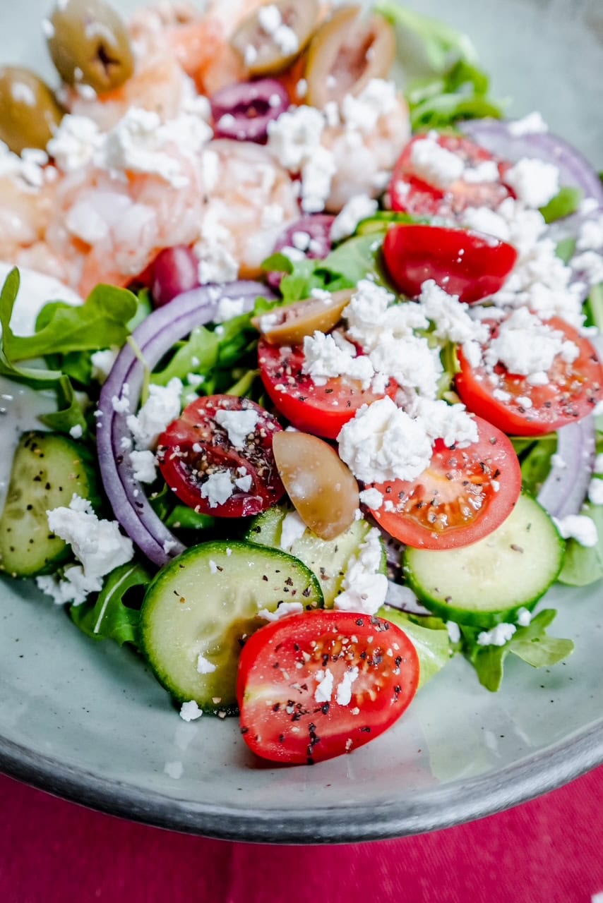 A fresh salad with cherry tomatoes, cucumber slices, red onion, green olives, arugula, crumbled feta cheese, and shrimp on a white plate. The salad is lightly seasoned with black pepper.