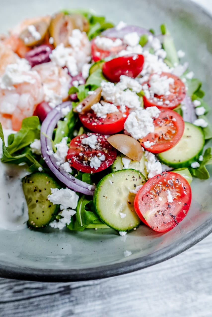 A close-up of a fresh salad with sliced cucumbers, cherry tomatoes, red onion, olives, mixed greens, and crumbled feta cheese in a bowl. Some pieces of shrimp and creamy dressing are visible on the side.
