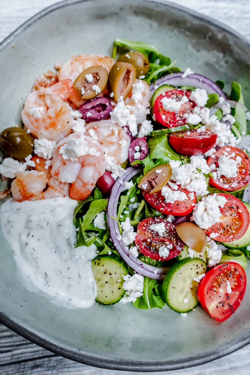 A bowl with shrimp, green olives, grape tomatoes, cucumber slices, red onion, arugula, crumbled feta cheese, and a dollop of creamy white sauce on the side.