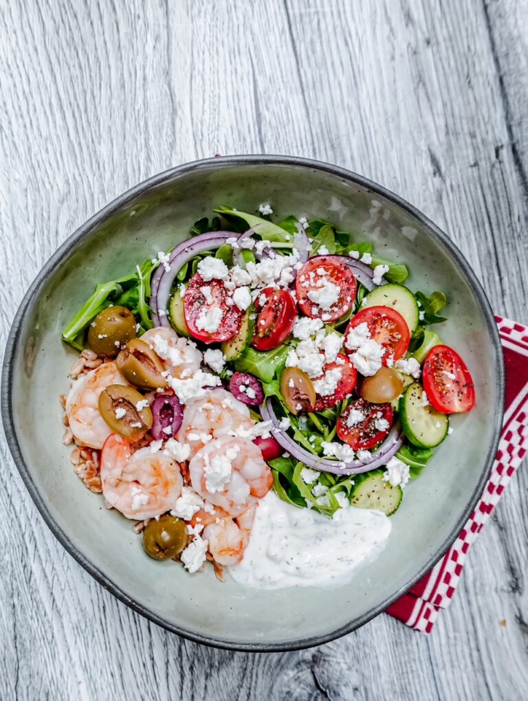 A bowl of salad with shrimp, cherry tomatoes, cucumber, olives, red onion, feta cheese, greens, and a dollop of creamy dressing, placed on a wooden surface with a red napkin underneath.