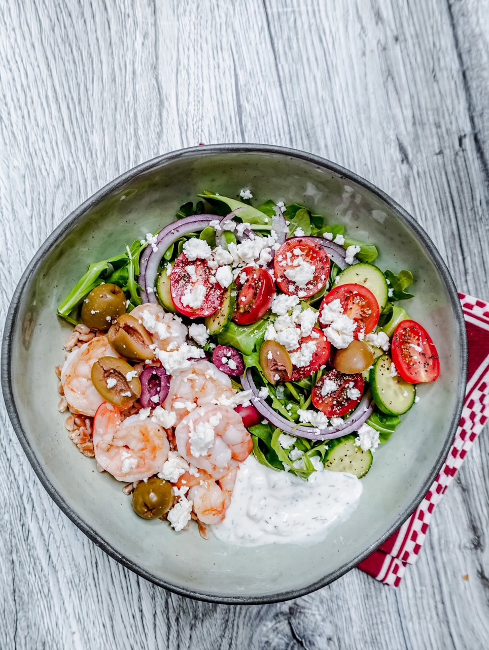 A bowl of salad with shrimp, cherry tomatoes, cucumber, olives, red onion, feta cheese, greens, and a dollop of creamy dressing, placed on a wooden surface with a red napkin underneath.