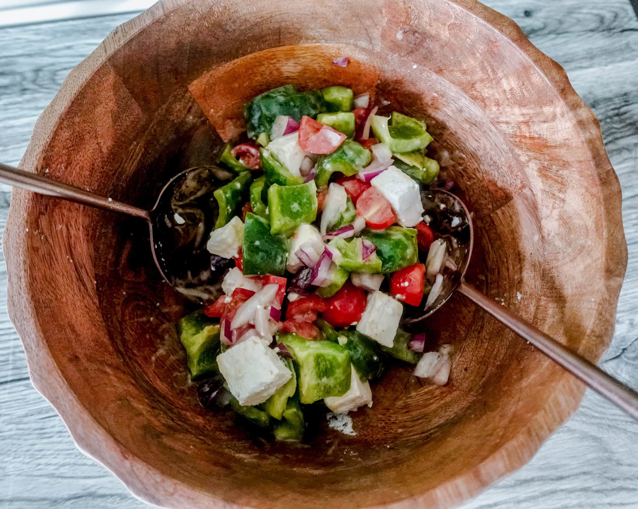 A wooden bowl filled with a fresh Greek salad containing green peppers, red onions, tomatoes, and feta cheese, with salad tongs on the sides.