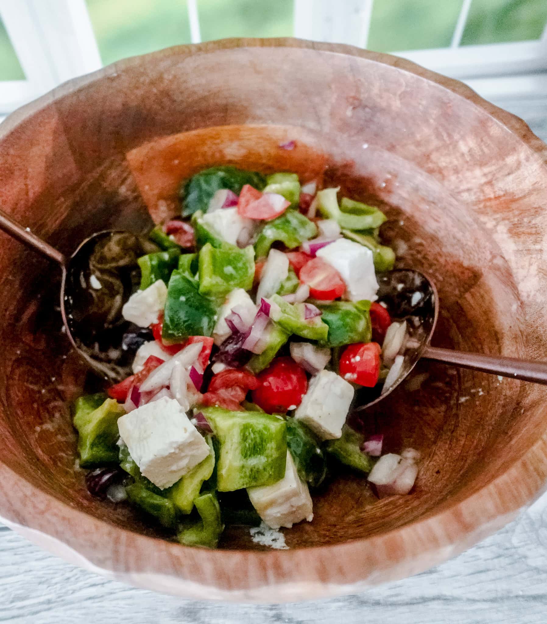 A wooden bowl filled with a fresh, colorful salad containing chopped green peppers, cherry tomatoes, red onion, and chunks of feta cheese.
