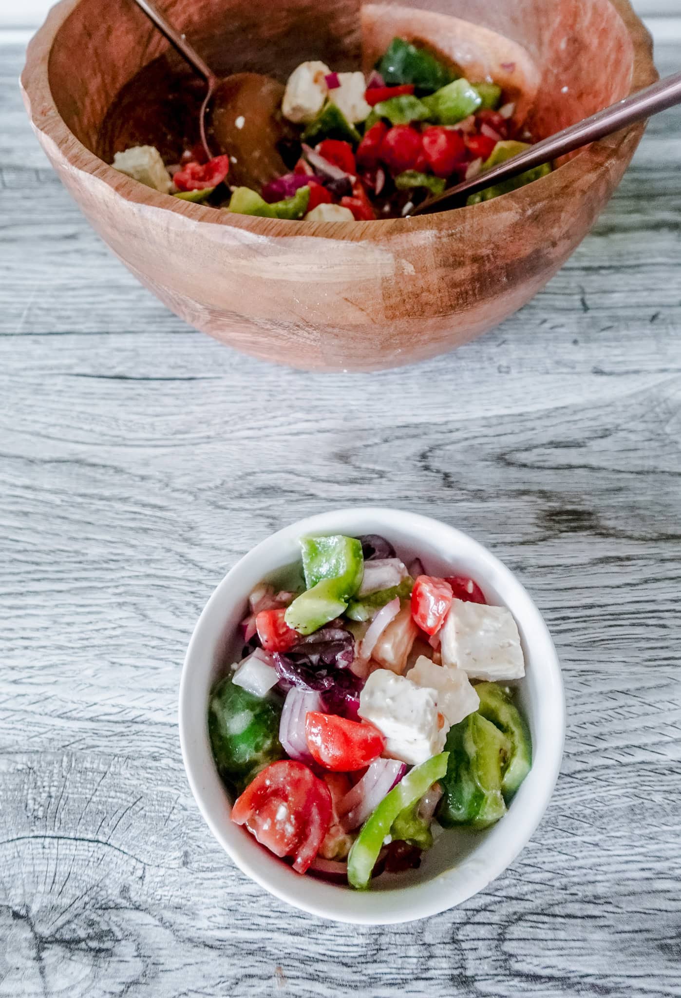A wooden bowl filled with vibrant Greek salad, featuring feta, tomatoes, olives, and green peppers. A smaller bowl in front displays a serving.