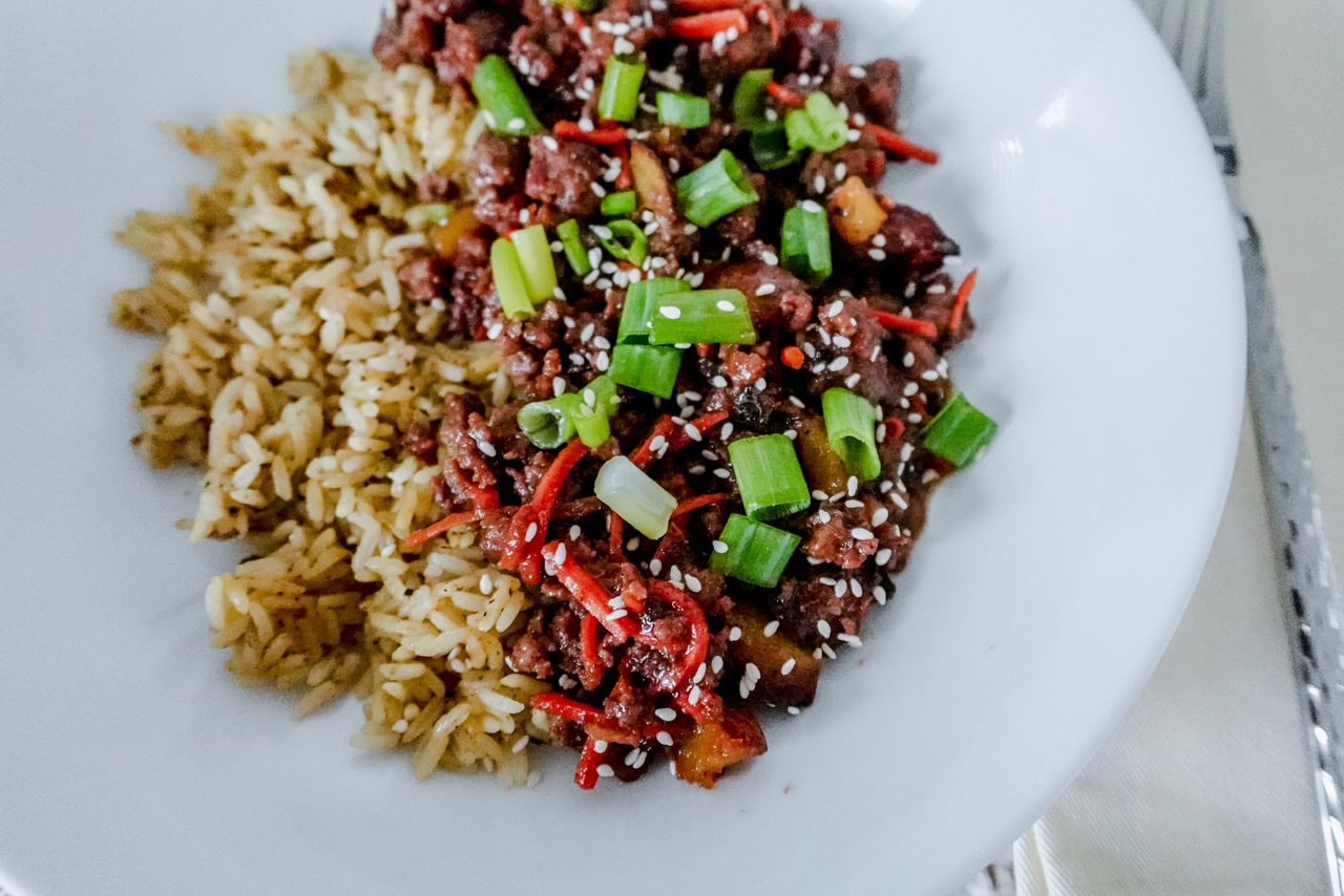 A white plate with seasoned rice and stir-fried beef garnished with potatoes, carrots, green onions, and sesame seeds. The dish looks vibrant and appetizing.