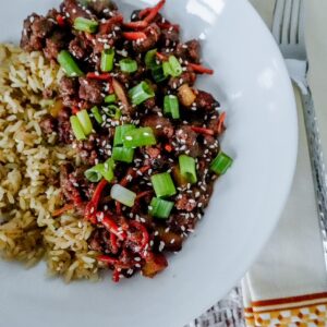 A white plate with stir-fried beef, carrots, and green onions garnished with sesame seeds, next to a serving of rice. A fork and napkin are on the side. Everything sits on a textured white tablecloth.
