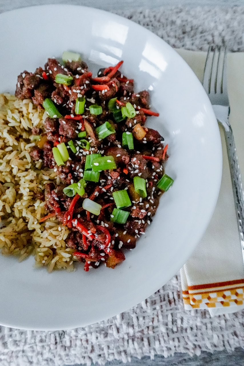 A white plate with stir-fried beef, carrots, and green onions garnished with sesame seeds, next to a serving of rice. A fork and napkin are on the side. Everything sits on a textured white tablecloth.