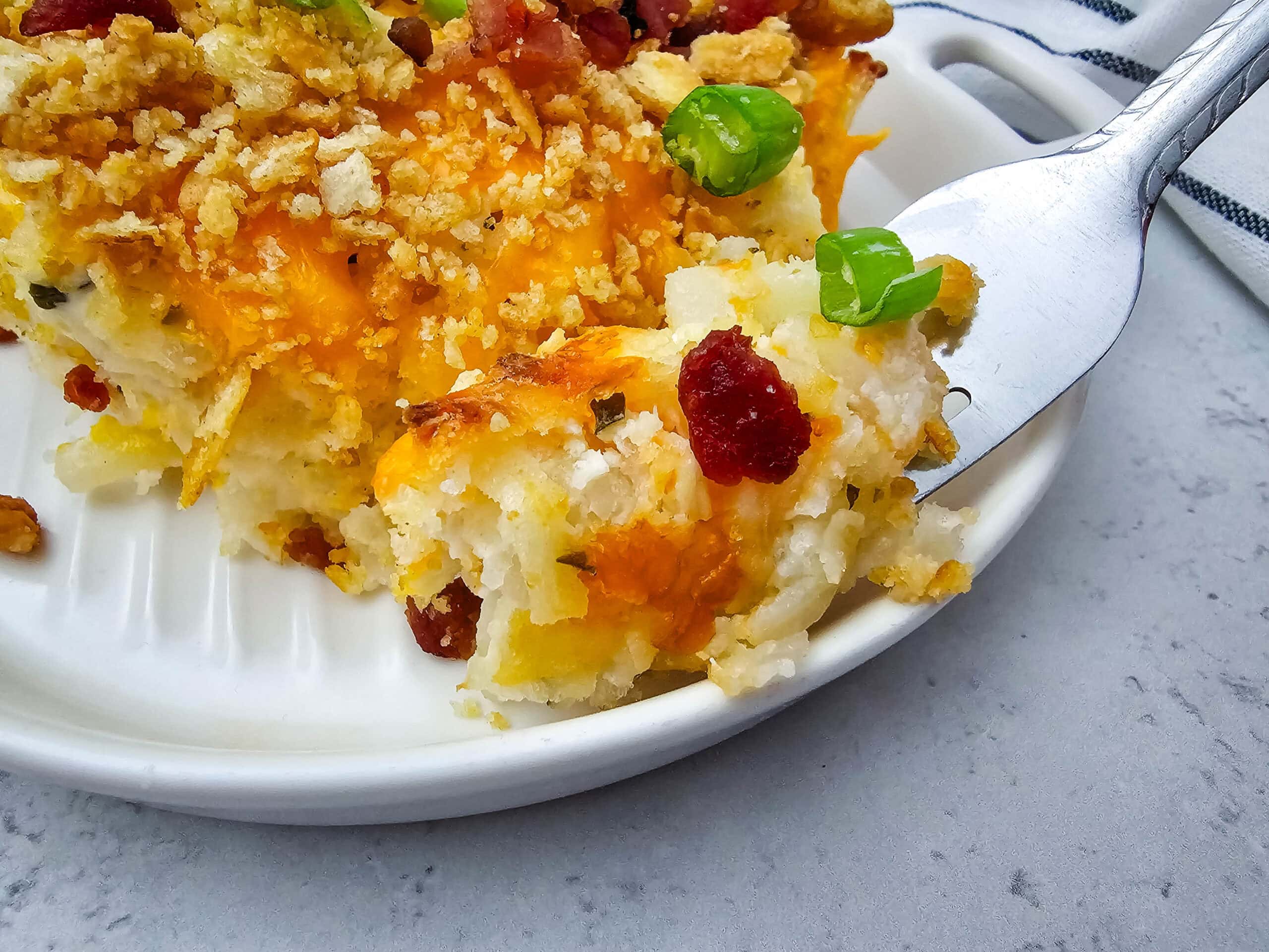 A close-up of a creamy, cheesy potato casserole that is topped with crispy bacon, golden crumbled ritz crackers and green onions.