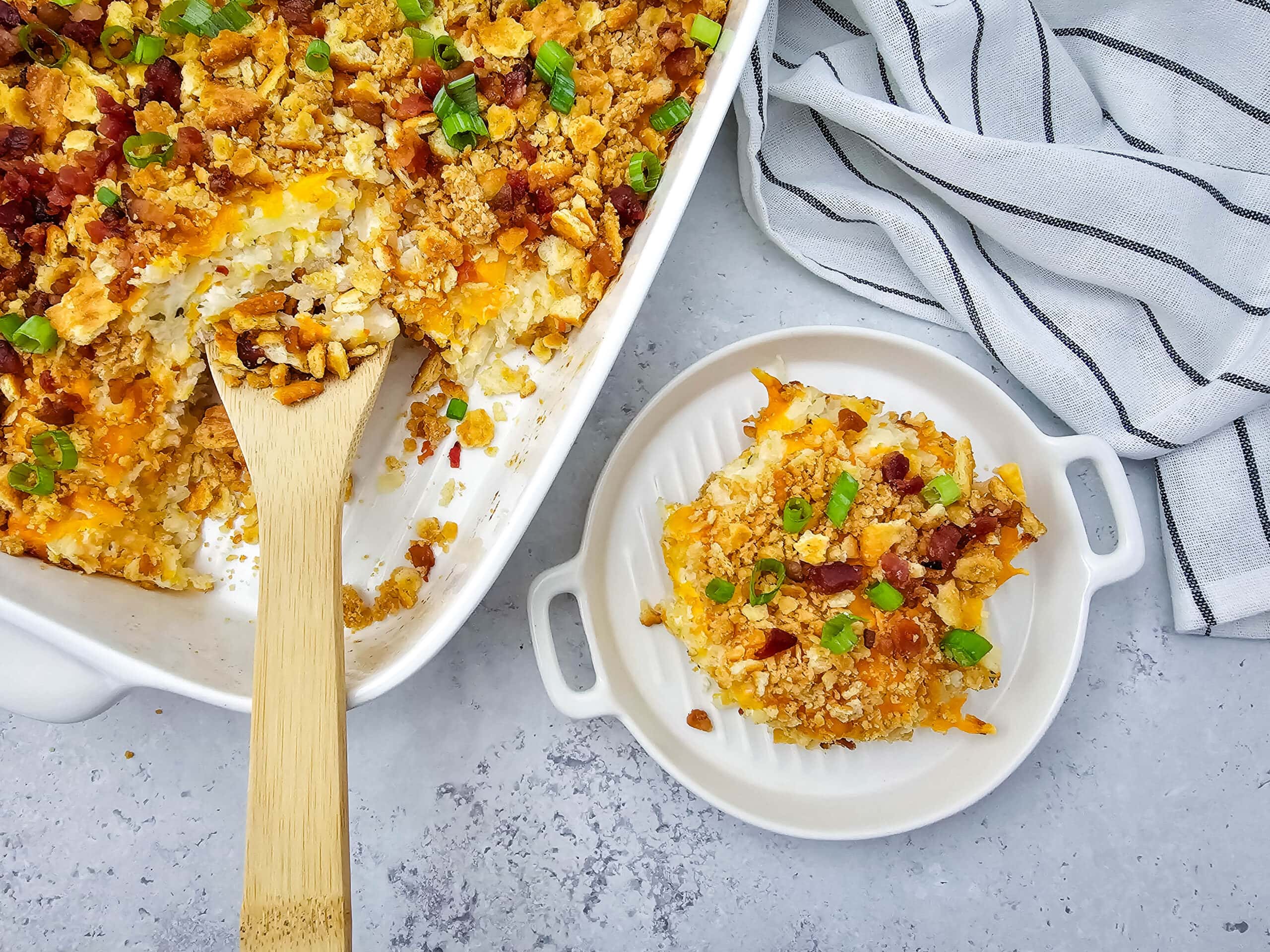 A large baking dish is filled with a creamy, cheesy potato casserole topped with crispy bacon, golden crumbled ritz crackers and green onions. A single serving sits on a smaller white dish next to the bigger dish. Everything sits on a gray/white countertop with striped towel in the background.