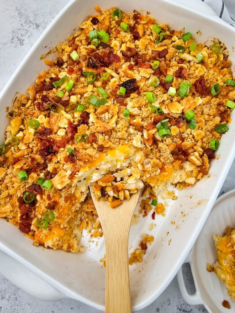 A wooden spoon rests in a large baking dish filled with a creamy, cheesy potato casserole topped with crispy bacon, golden crumbled ritz crackers and green onions.