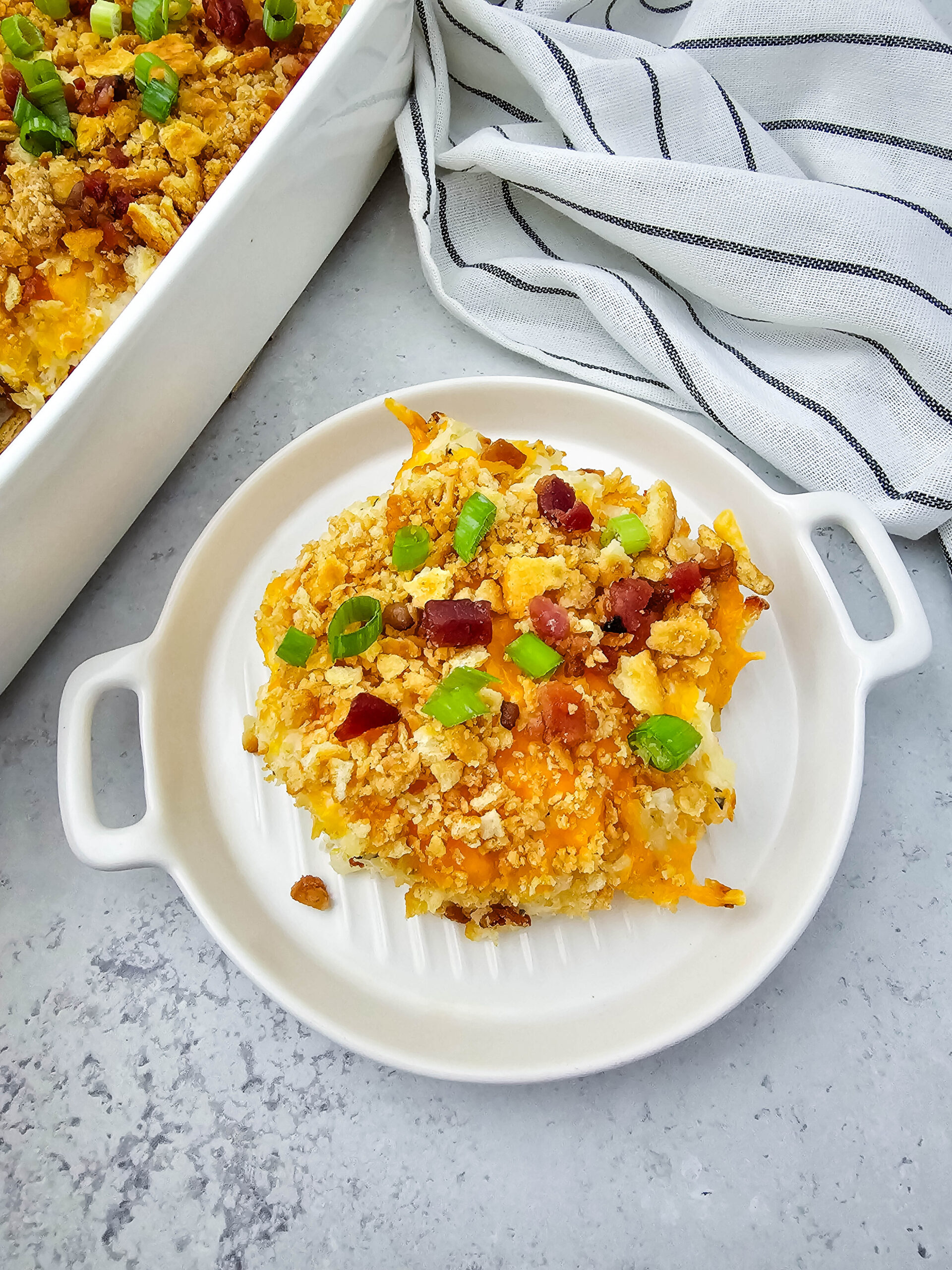 A single serving of creamy, cheesy potato casserole sits on a small white dish, topped with golden Ritz cracker crumbs, crispy bacon, and fresh green onions. Behind it, a larger baking dish with the rest of the casserole and a wooden spoon adds a cozy, homemade touch on a gray-and-white countertop with a striped towel in the background.