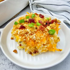 A single serving of creamy, cheesy potato casserole sits on a small white dish, topped with golden Ritz cracker crumbs, crispy bacon, and fresh green onions. Behind it, a larger baking dish with the rest of the casserole and a wooden spoon adds a cozy, homemade touch on a gray-and-white countertop with a striped towel in the background.