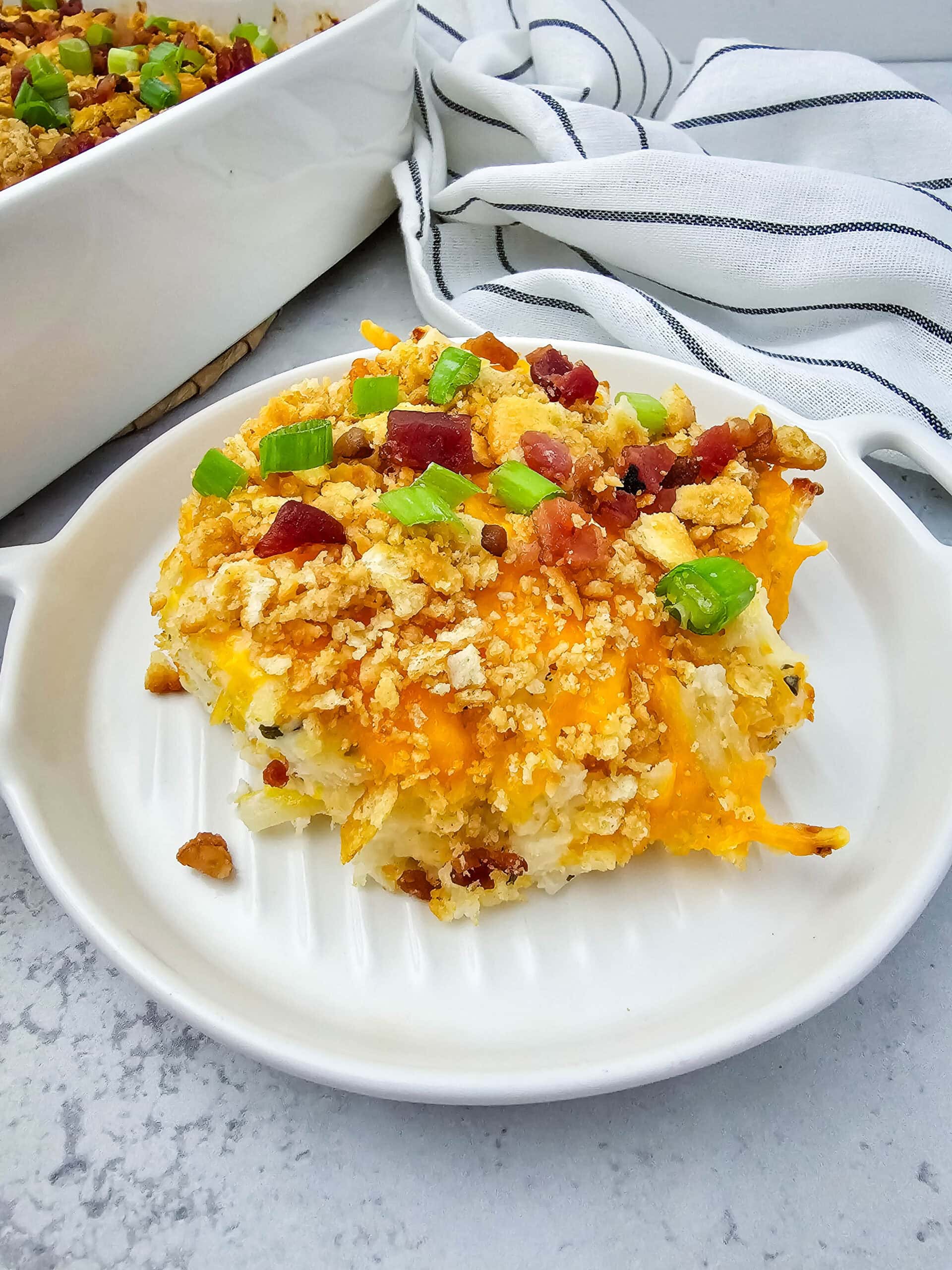 A single serving of creamy, cheesy potato casserole sits on a small white dish, topped with golden Ritz cracker crumbs, crispy bacon, and fresh green onions. Behind it, a larger baking dish with the rest of the casserole and a wooden spoon adds a cozy, homemade touch on a gray-and-white countertop with a striped towel in the background.