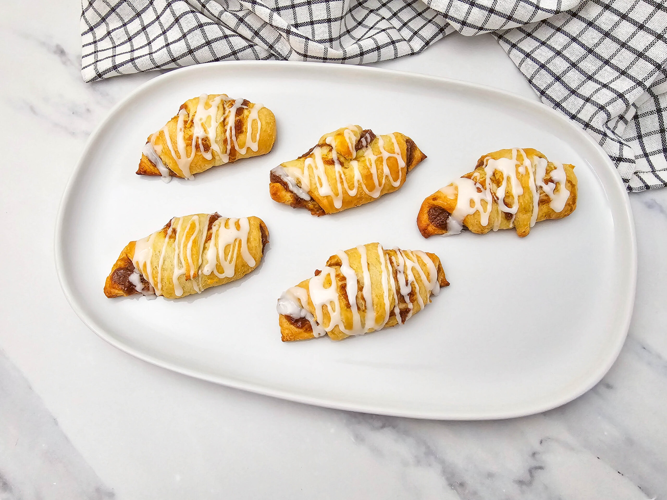 Five pumpkin crescent rolls with a cinnamon filling, drizzled with white icing, are arranged on a white plate. A checkered kitchen towel is in the background.