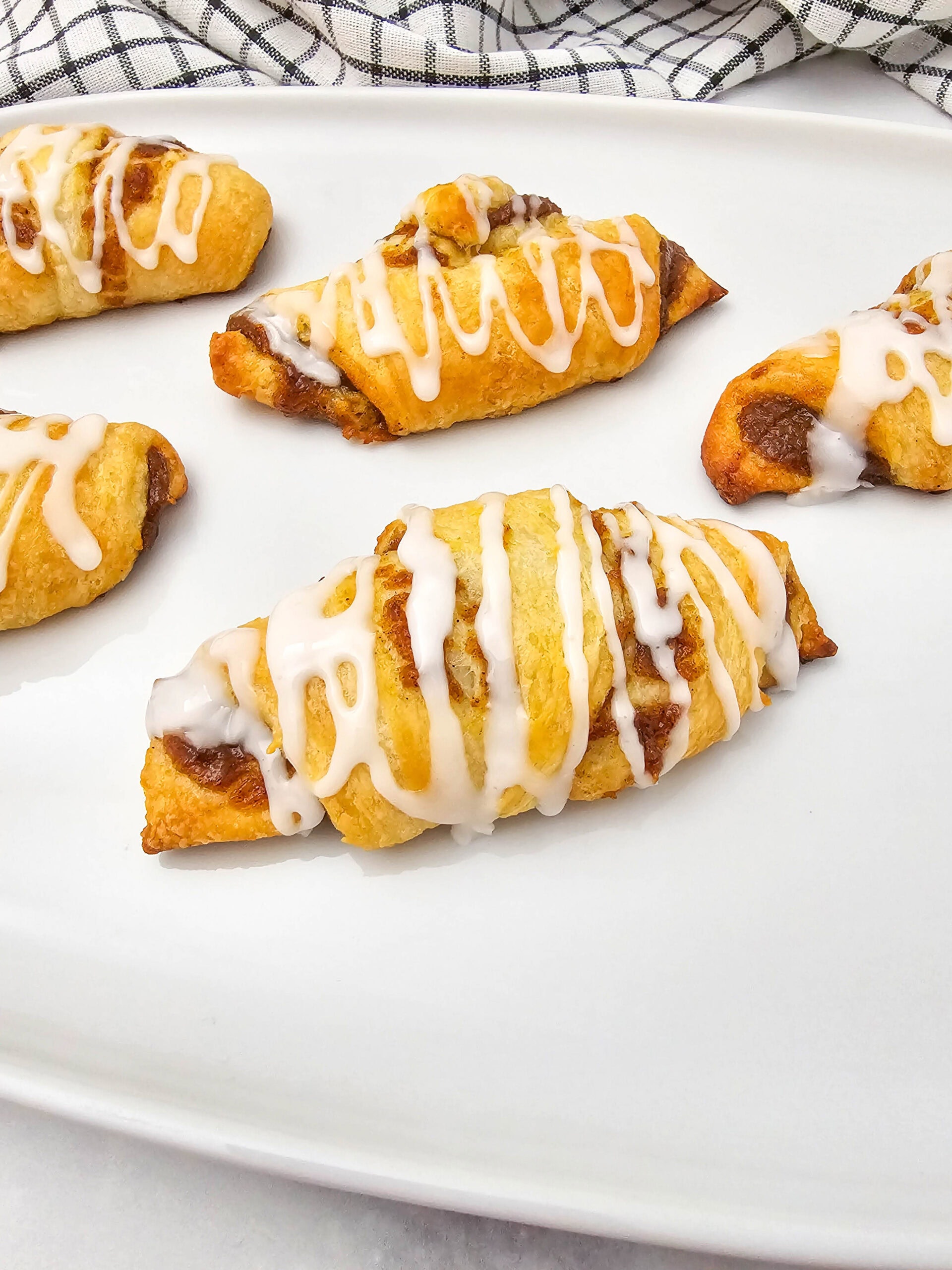 Five crescent-shaped pumpkin pastries with white icing drizzle are arranged on a white plate. A checkered cloth lies in the background.