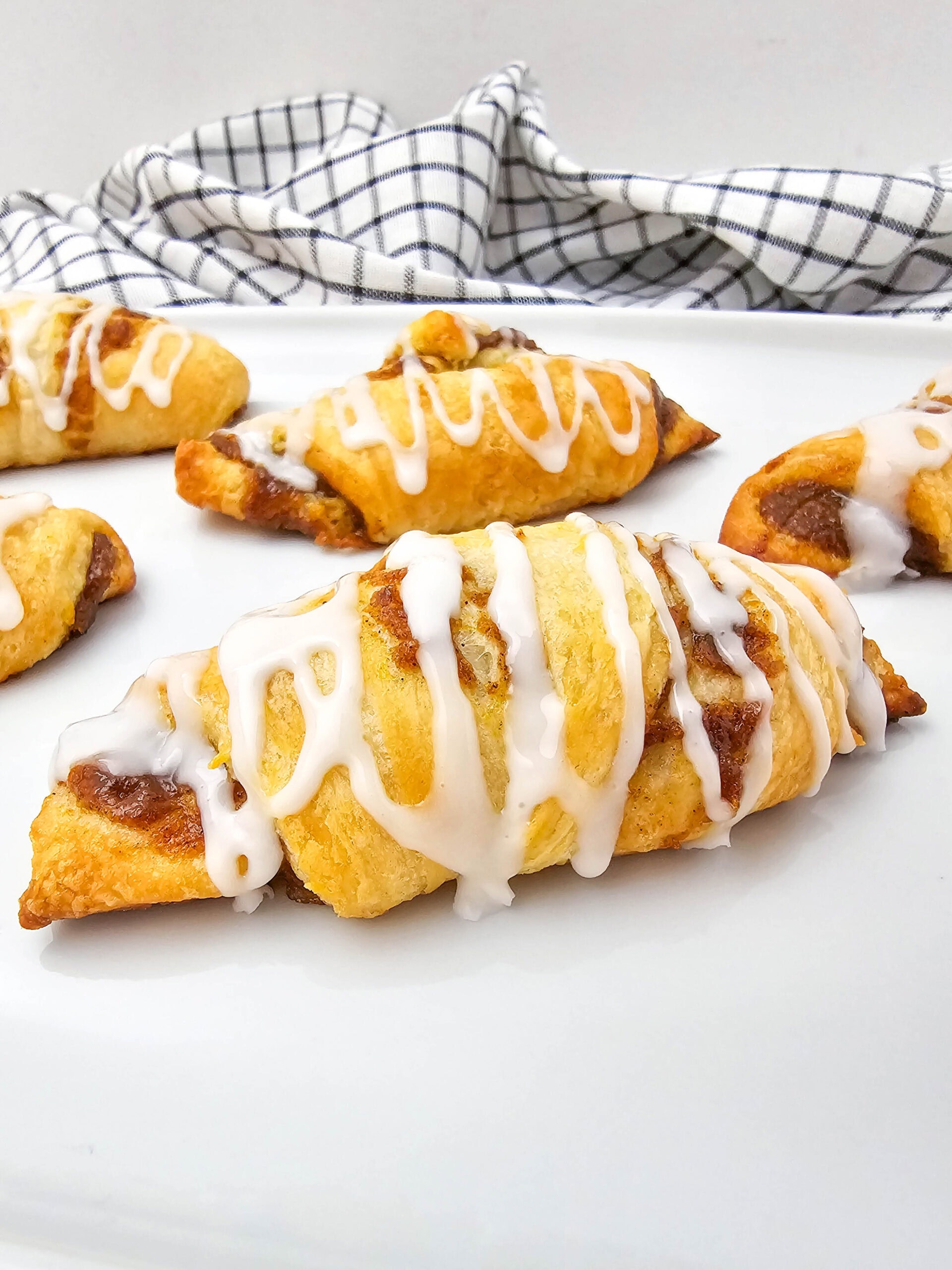 Golden-brown pumpkin crescent rolls drizzled with white icing are displayed on a white surface, with a black-and-white checkered cloth in the background.