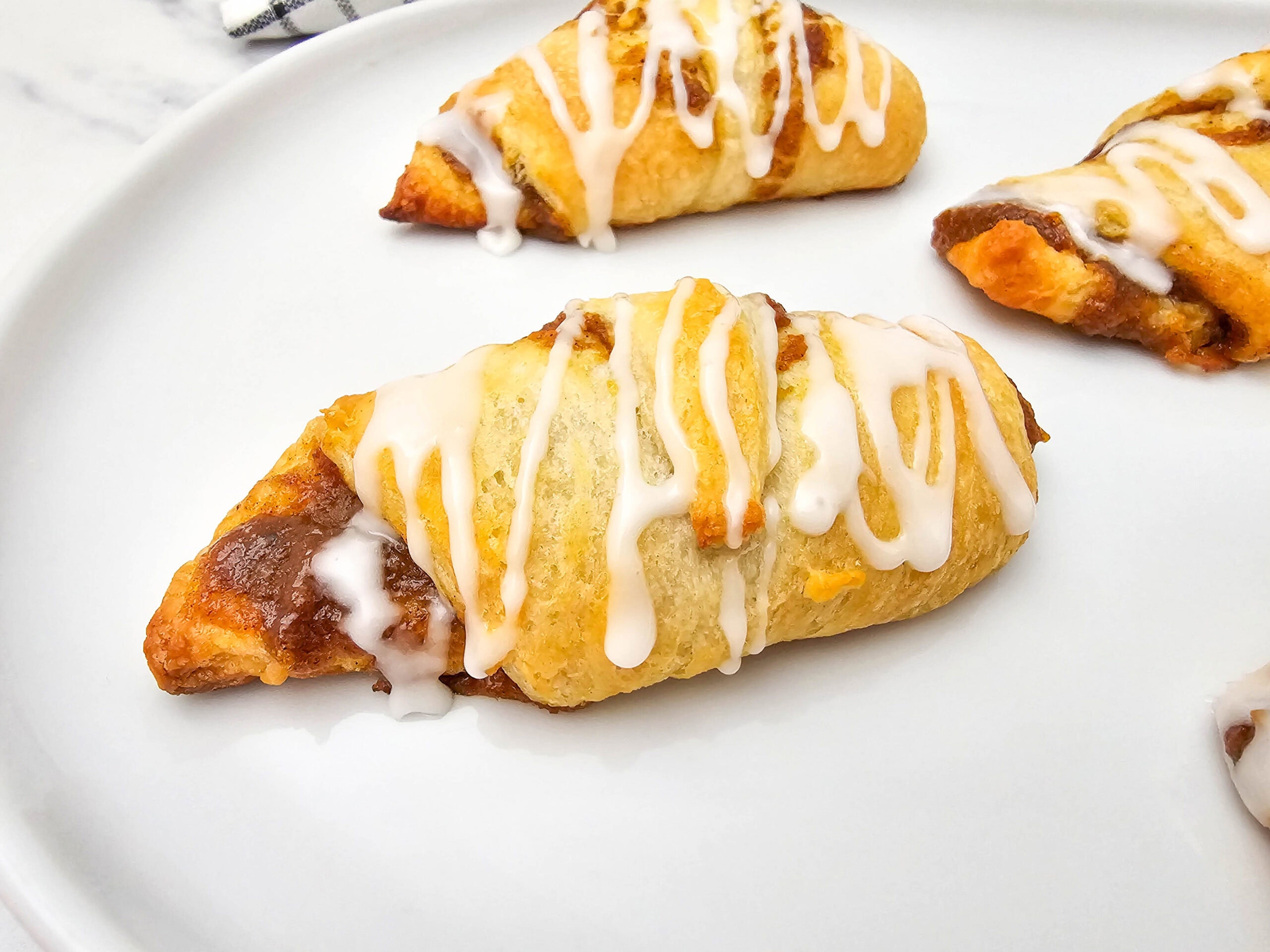 A close-up of pumpkin crescent rolls drizzled with white icing, arranged on a white plate. The rolls appear golden brown and flaky, with visible layers and a sweet filling inside.