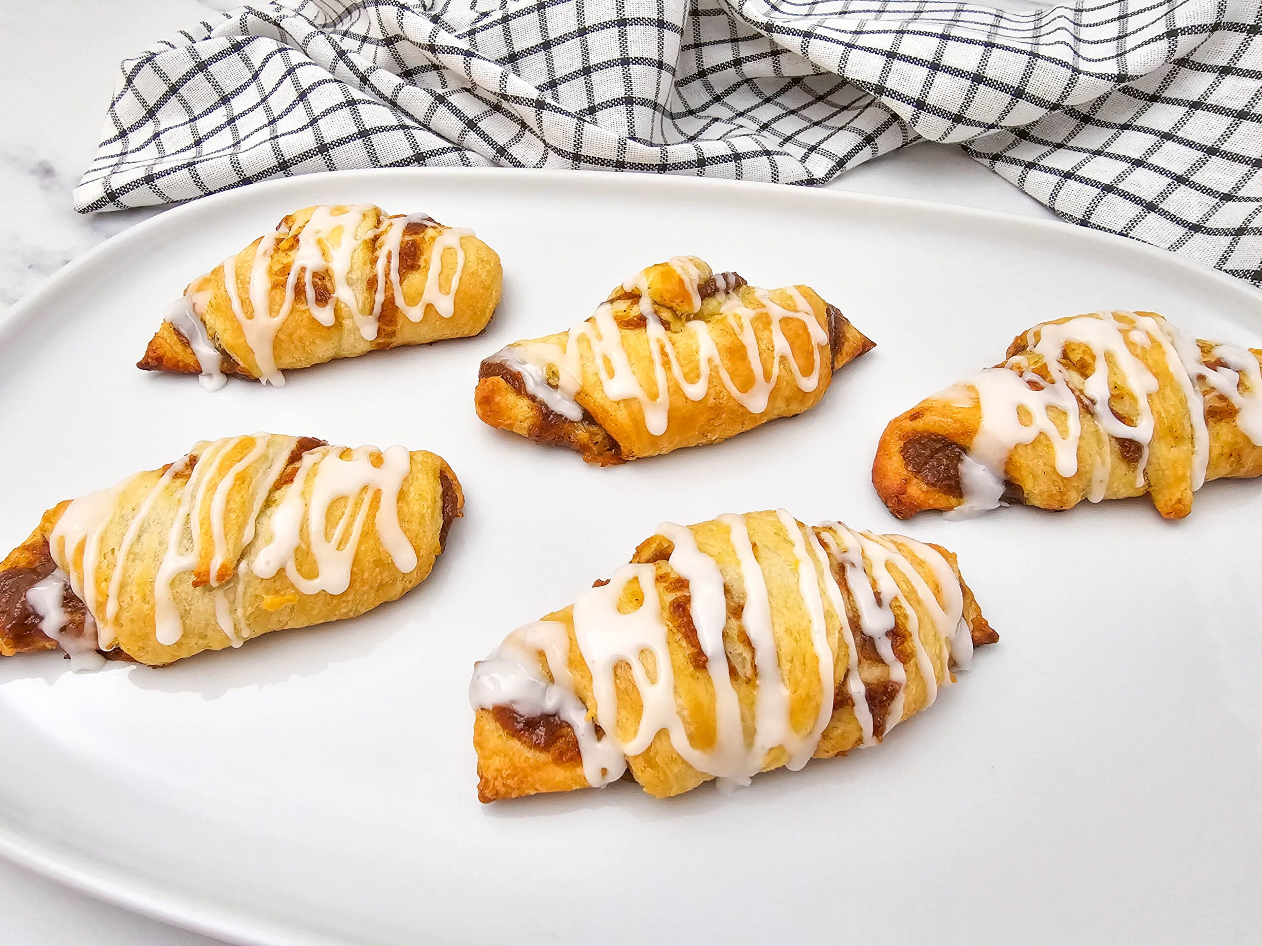Five pumpkin crescent rolls with a cinnamon filling, drizzled with white icing, are arranged on a white plate. A checkered kitchen towel is in the background.